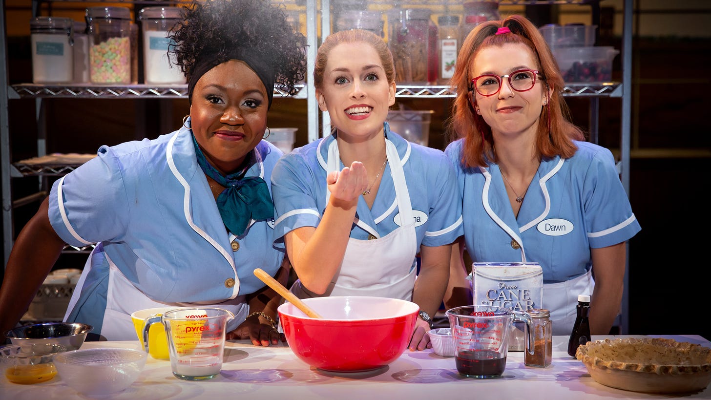 Kennedy Salters as Becky, Bailey McCall as Jenna, and Gabriella Marzetta as Dawn lean over a mixing bowl on a kitchen counter top dressed in waitress uniforms.