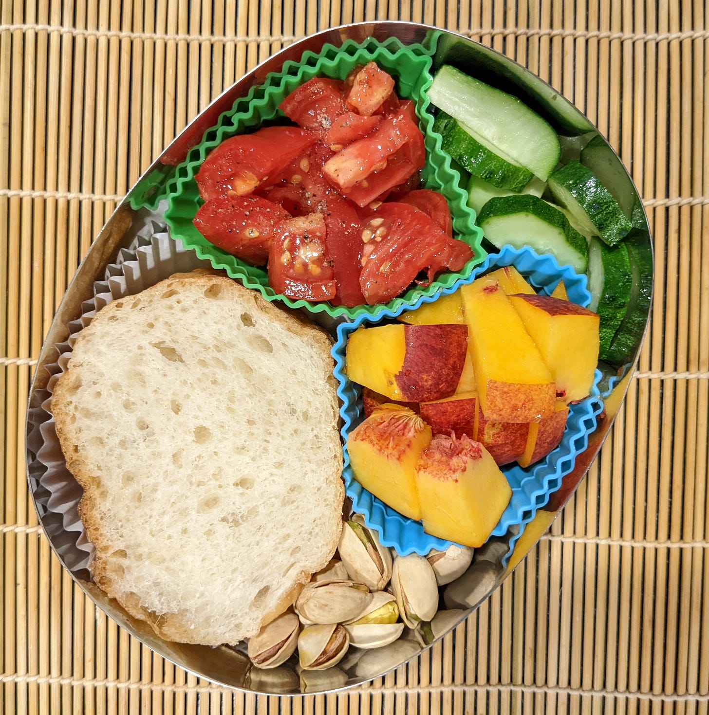A metal bento box filled with fresh tomatoes, cucumber, nectarines, pistachios, and bread