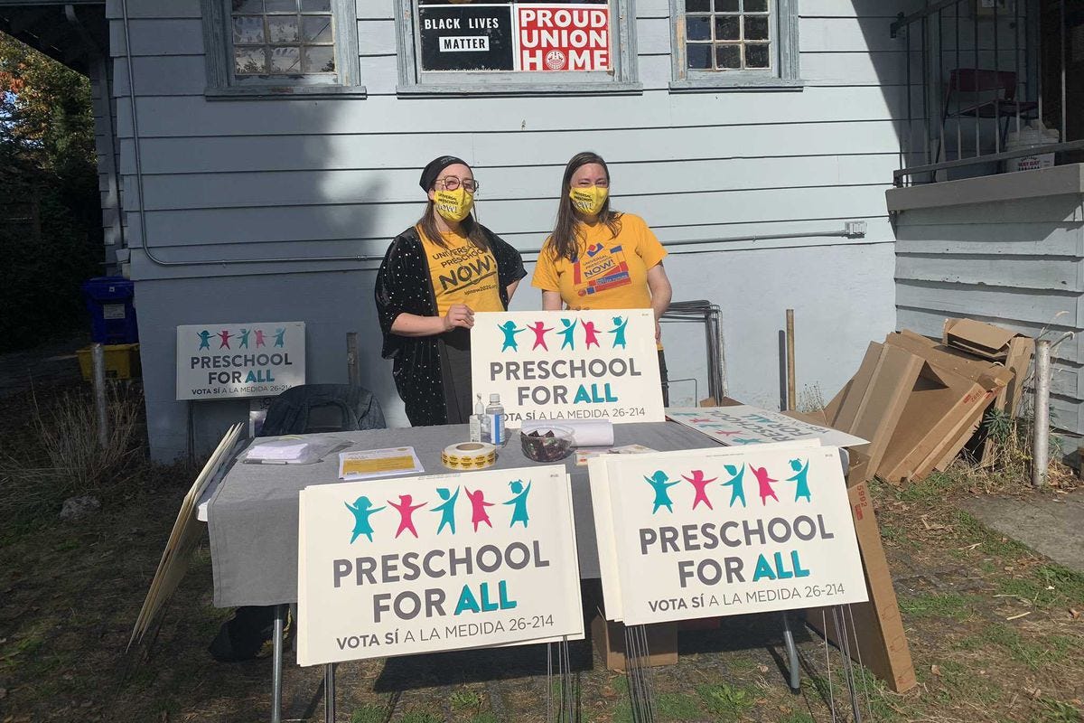 Two masked volunteers hand out signs reading “Preschool for All.”