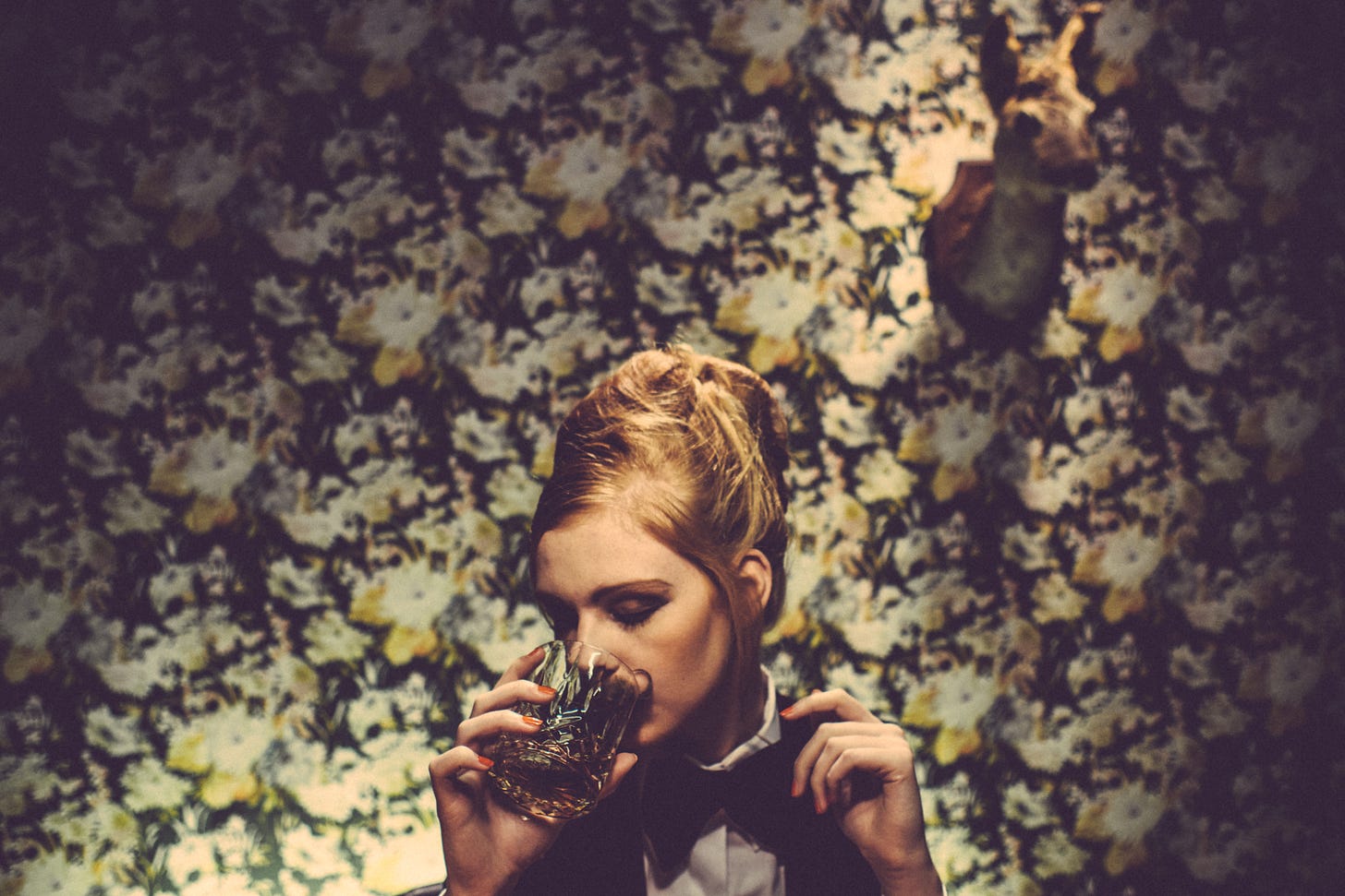 Woman with an updo drinking a cocktail in front of garish wallpaper with a taxidermied head of some sort on the wall