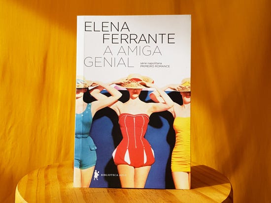 Descrição: Foto do livro A Amiga Genial, de Elena Ferrante, sobre uma banqueta de madeira em frente a um fundo amarelo. A capa do livro traz a imagem de três mulheres banhistas com maiôs dos anos 40 e o título na parte de cima.