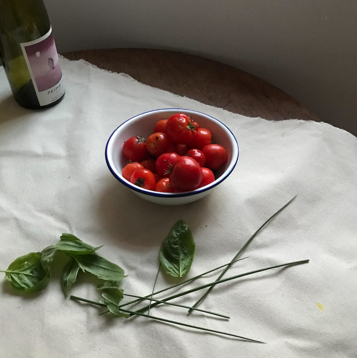 A piece of cream canvas is spread at an angle across a table. To the left, a large empty green glass wine bottle. In the centre is a white enamel bowl with a blue rim, full of bright red ripe tomatoes, of varying shapes and sizes. They gleam. In the foreground, the split long stems and leaves of some basil. 