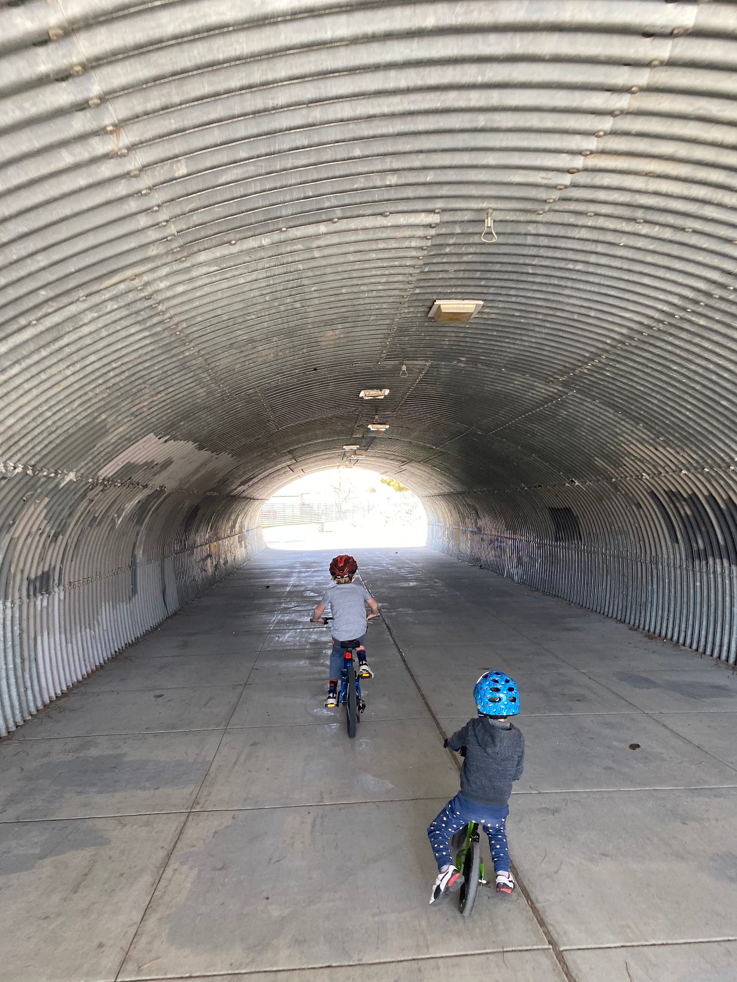 My two kids (one smaller than the other), riding their bikes through a corrugated metal tunnel, towards the sunlight