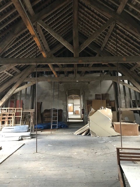 Huge attic with vaulted ceiling at the top of an old convent in france. the space has piles of old furniture and boxes, and there are many wooden beams. It's sunny.