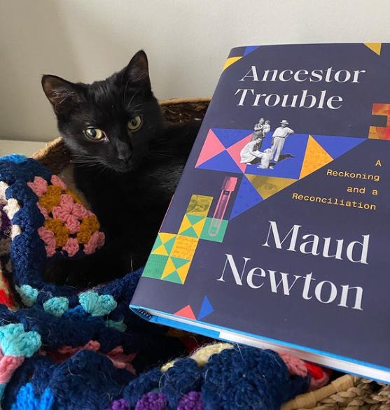 Image shows a black cat on a multicolored afghan in a basket, with Maud Newton's Ancestor Trouble visible in the foreground.