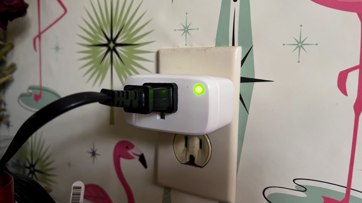 An Eve Energy smart plug with its light on and a black plug in it on a wallpapered backdrop