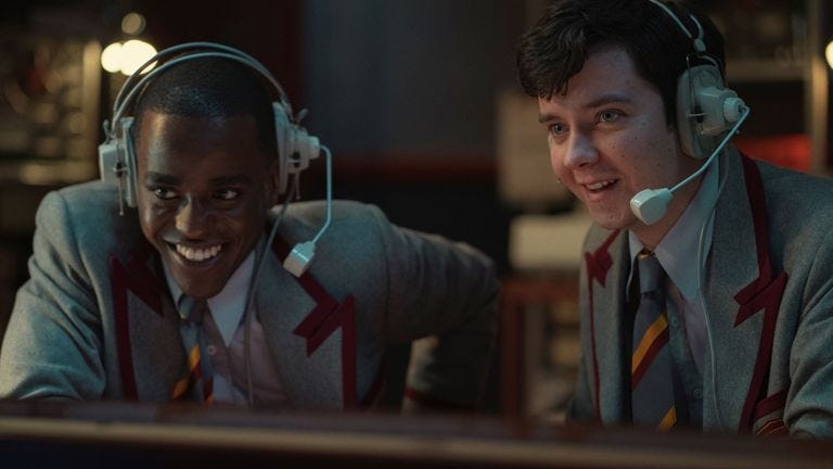 This image shows Ncuti Gatwa (left) as Eric Effiong and Asa Butterfield (right) as Otis Milburn in Laurie Nunn's Netflix series Sex Education. Both characters wear school jackets and ties, as well as headsets with microphones, while smiling and looking off camera. This image shows Ncuti Gatwa (left) as Eric Effiong and Asa Butterfield (right) as Otis Milburn in Laurie Nunn's Netflix series Sex Education. Both characters wear school jackets and ties, as well as headsets with microphones, while smiling and looking off camera.