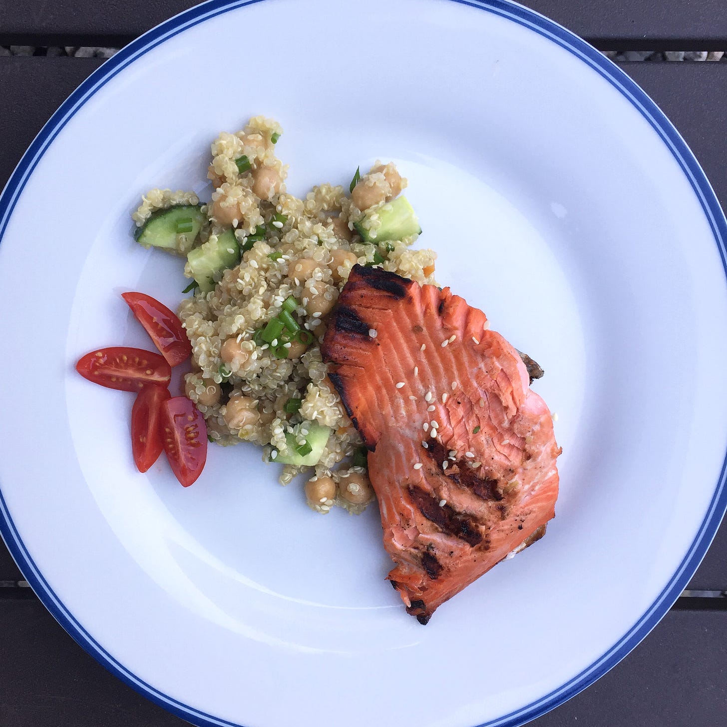 a white plate with quinoa and chickpea salad beneath a piece of grilled dark pink salmon. Cucumbers and sesame seeds are visible in the salad, and a sliced grape tomato sits to the side.
