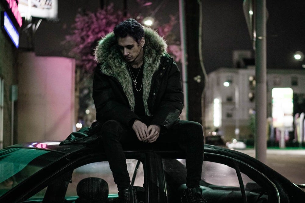cøzybøy in a coat with a large faux fur collar looking down. He is sitting on top of a car outside.
