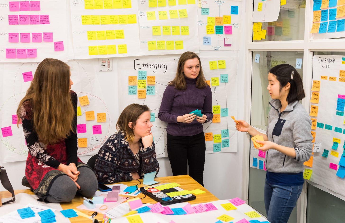 Four Olin students stand in a design studio discussing a project, surrounded by posters and graphs and dozens of sticky notes, processing ideas.