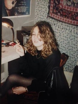 Picture of Kathryn Mockler, a white teenager with brown curly hair sitting at a white desk with her eyes closed holding a cigarette. She wears all black. A white and black patterned scarf hangs on the wall beside a Pink Floyd poster. 