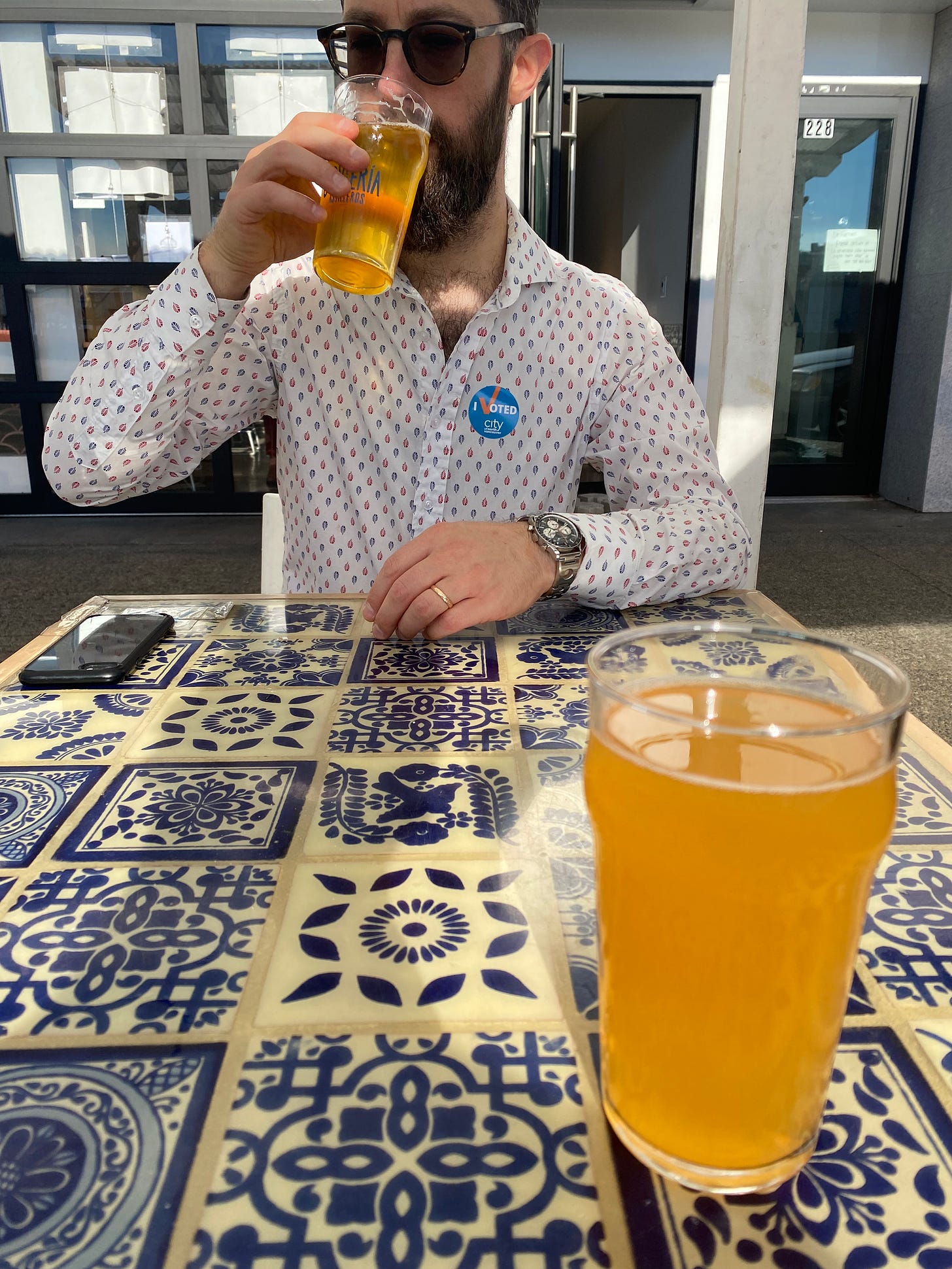 A table made of white and blue Mexican tiles, with a golden glass of beer in the foreground. Across the table, Jeff sips his own glass of beer. He is wearing sunglasses and a white shirt with a blue and arrow pattern, and there is a blue "I voted" sticker on his chest.