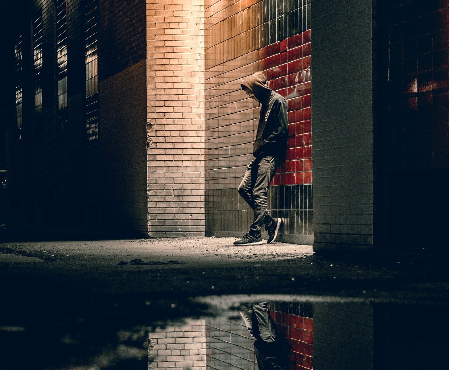 man wearing hooded sweatshirt leaning against brick building at night under spotlight