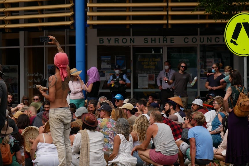 A large group of people without masks sitting on the ground. Police in background.