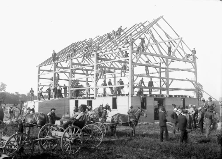 Barn raising, called Leckie’s Barn. Horse-drawn carts in front of a completed barn frame, men leaning and standing in rafters