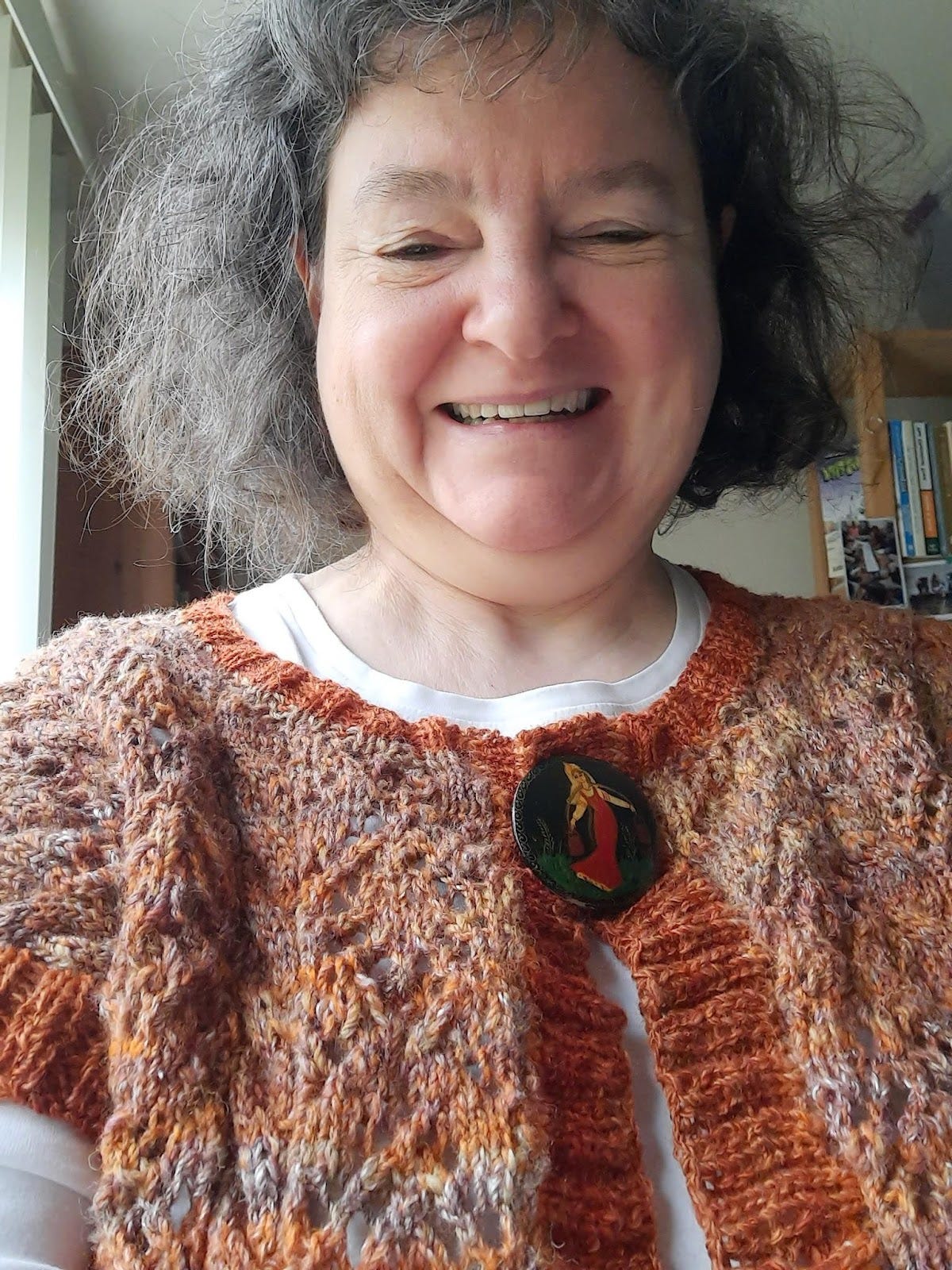 A dark haired white woman grins into the camera. She is wearing a handknitted lace top in rusty colors. She just finished knitting this top from her own handspun wool.