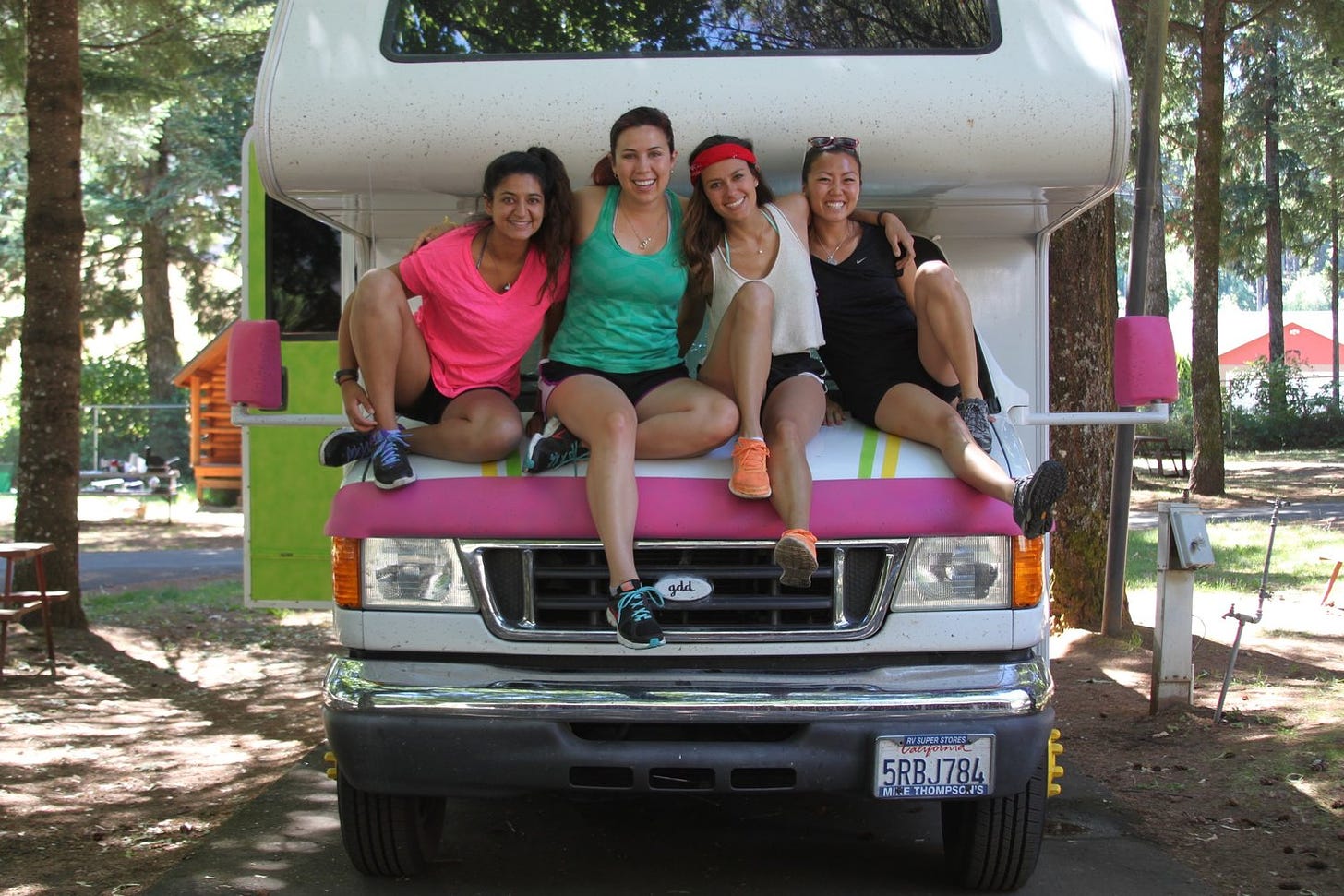 Katie is sitting on top of a large white and pink RV with three of her friends. They are all dressed in sports gear, smiling into the camera with their arms around each other. The RV is parked in between tall trees. The sun is shining.