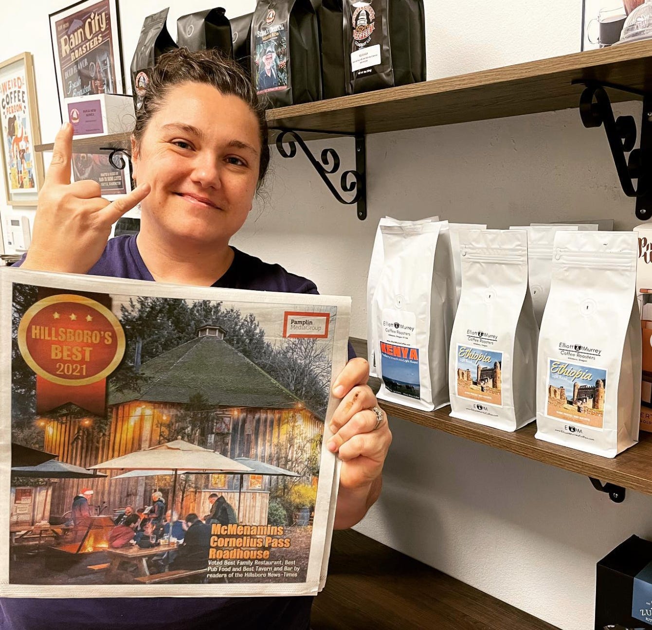 Keri Elliot flashes the rock n'roll horns hand signal while holding up a newspaper promoting her coffeeshops bronze medal for best coffeehouse in Hillsboro, Oregon while standing in from of shelves filled with coffee bags.