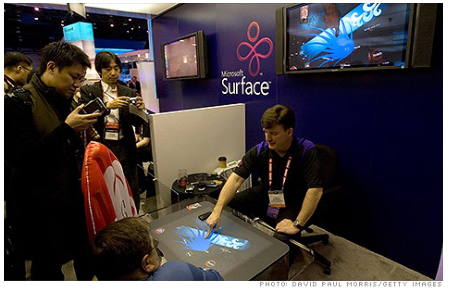 Microsoft employee at a trade show demonstrating the original Surface Table. He's seated in a standard chair touching the big screen that is on top of a table while people look on.