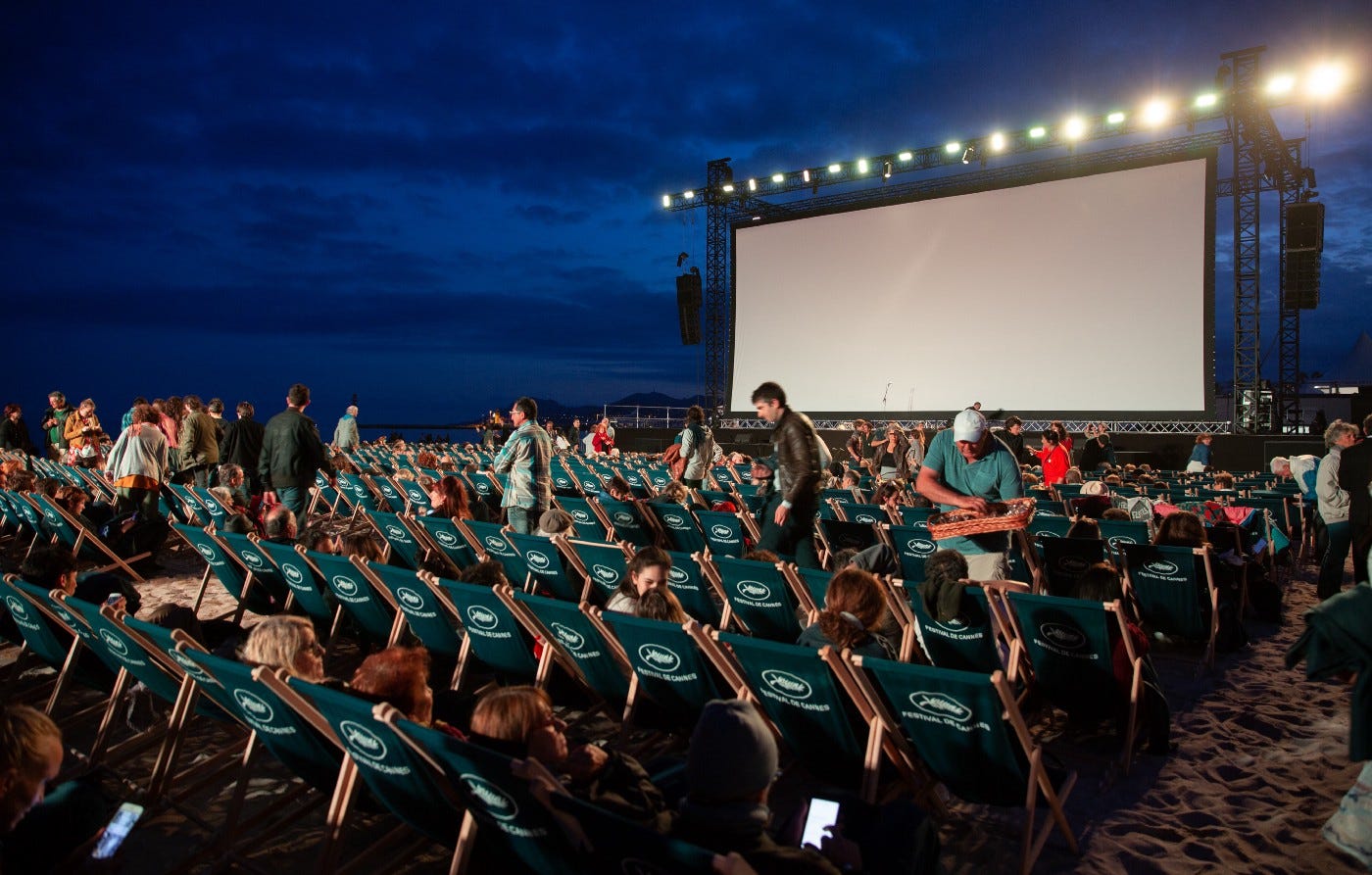 People sitting in seats in front of a large projector outdoors. The lights are on and some people are preparing stuff (moving around, buying food), while others have settled into their chairs and are awaiting the movie.