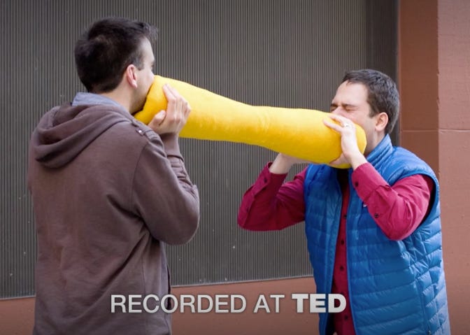 Two men scream into opposite ends of a large yellow tube
