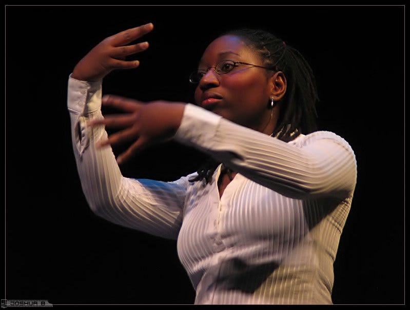 This is a photo of a Black woman wearing a white shirt, seeing-eye glasses, with twists in her hair as she interprets American Sign Language with her two hands in the air against a solid black background. 