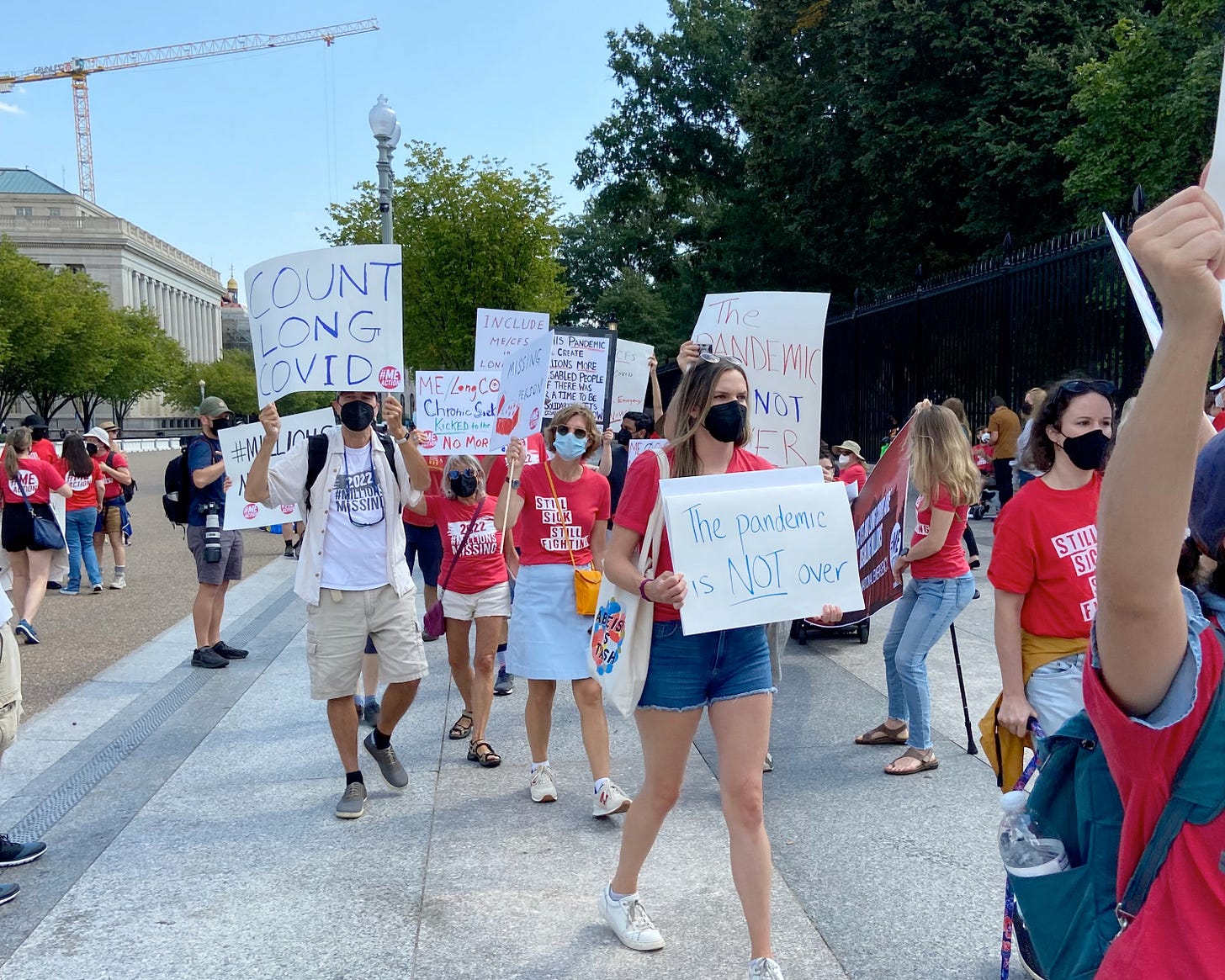 Long COVID ME/CFS protest at the White House: people on a sidewalk wearing masks and carrying signs that say the pandemic is not over and count long covid and missing person.