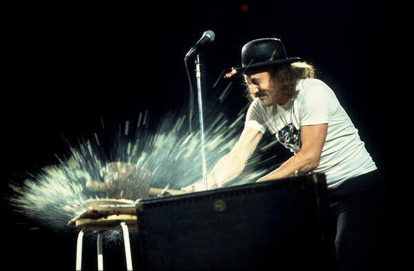 Gallagher, wearing a black hat, a white T-shirt, shoulder-length brown hair and a mustache, taking a sledgehammer to a large fruit during a performance.