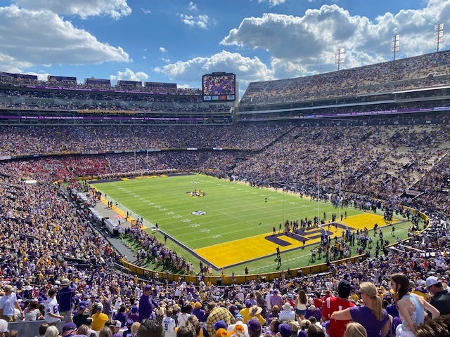 A near-capacity crowd fills the stands of LSU's Tiger Stadium, a massive college football stadium on a sunny day.
