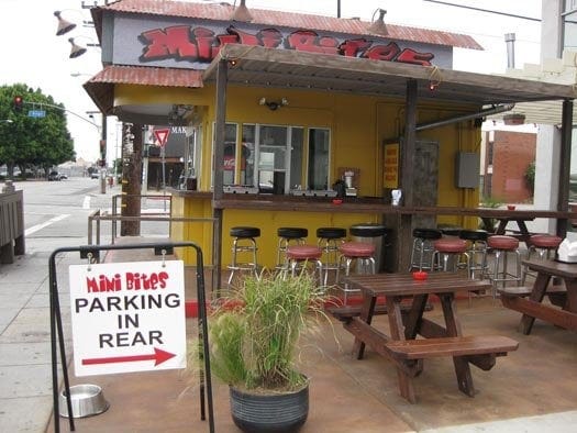 The shack is painted yellow, with a red sign that says Mini Bites and a metal sign in the front that indicates parking in the rear. There are brown picnic tables and a few barstools. There are no people present in the image.