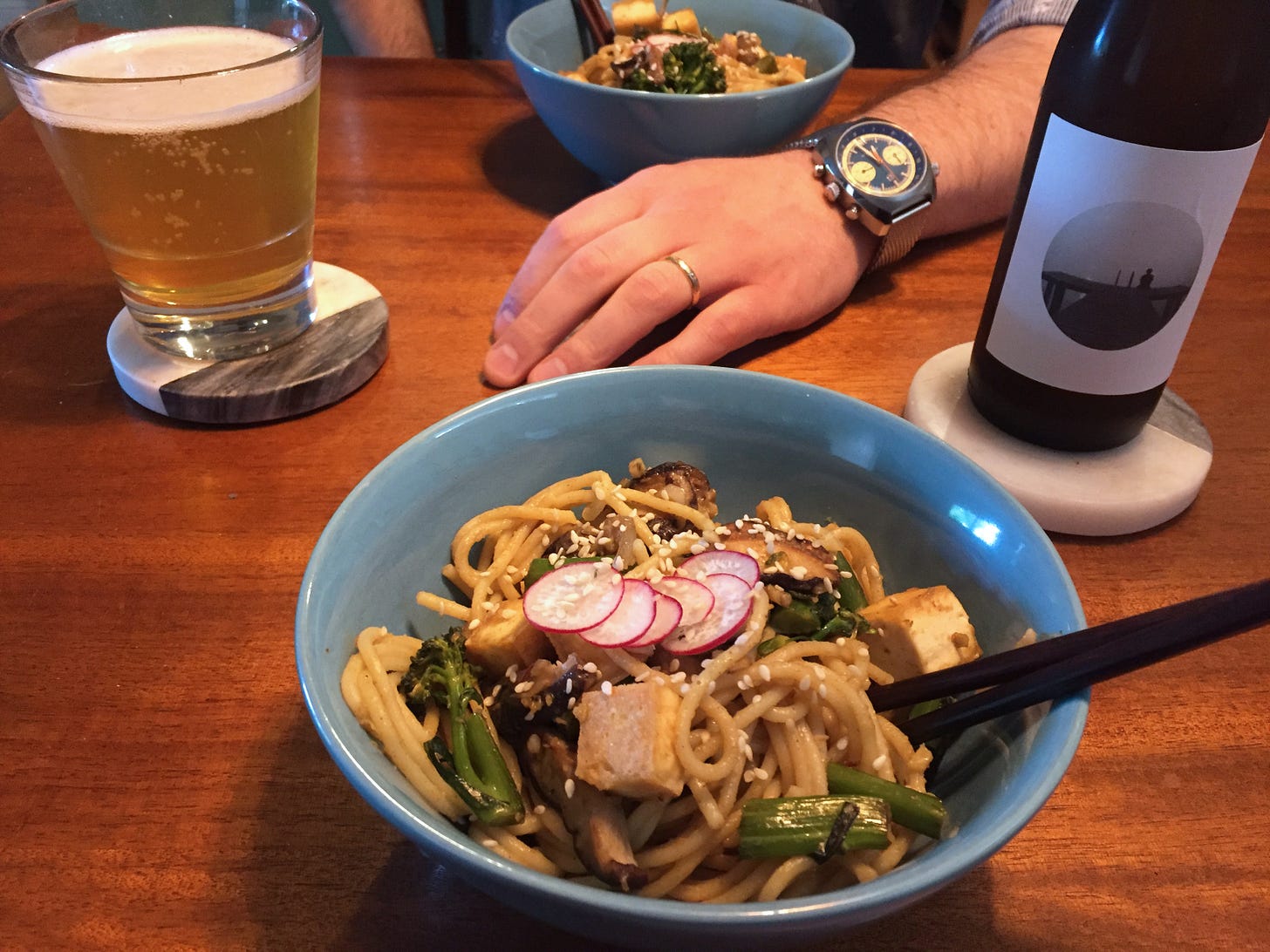 two blue bowls full of curried noodles with squares of tofu, pieces of broccolini, and mushroom slices. On top are sesame seeds and slices of red radish. A bottle of beer and a glass of beer sit at the respective corners of the bowls.