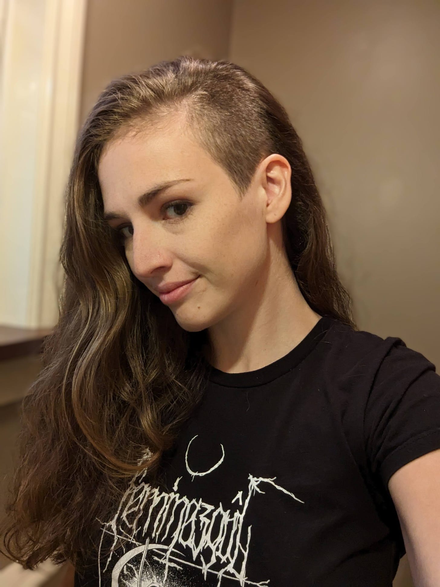 Molly White, a white woman with long brown hair that's shaved on one side. She's wearing a black t-shirt with the logo of the band Feminazgul on it in white.