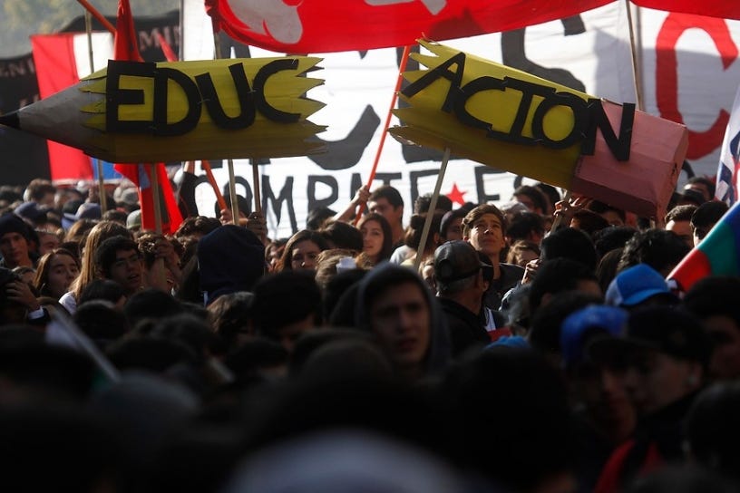 Another photo of a protest, this one is darker and more carefully composed. Marching heads bob in the foreground in shadow, while in the mid-ground, a group of students in a ray of light hold up a large cardboard pencil, broken in two down the middle. Written across the pencil in black block letters is the word "Educación," education, also split in two. Behind them, more students carry big banners and posters.