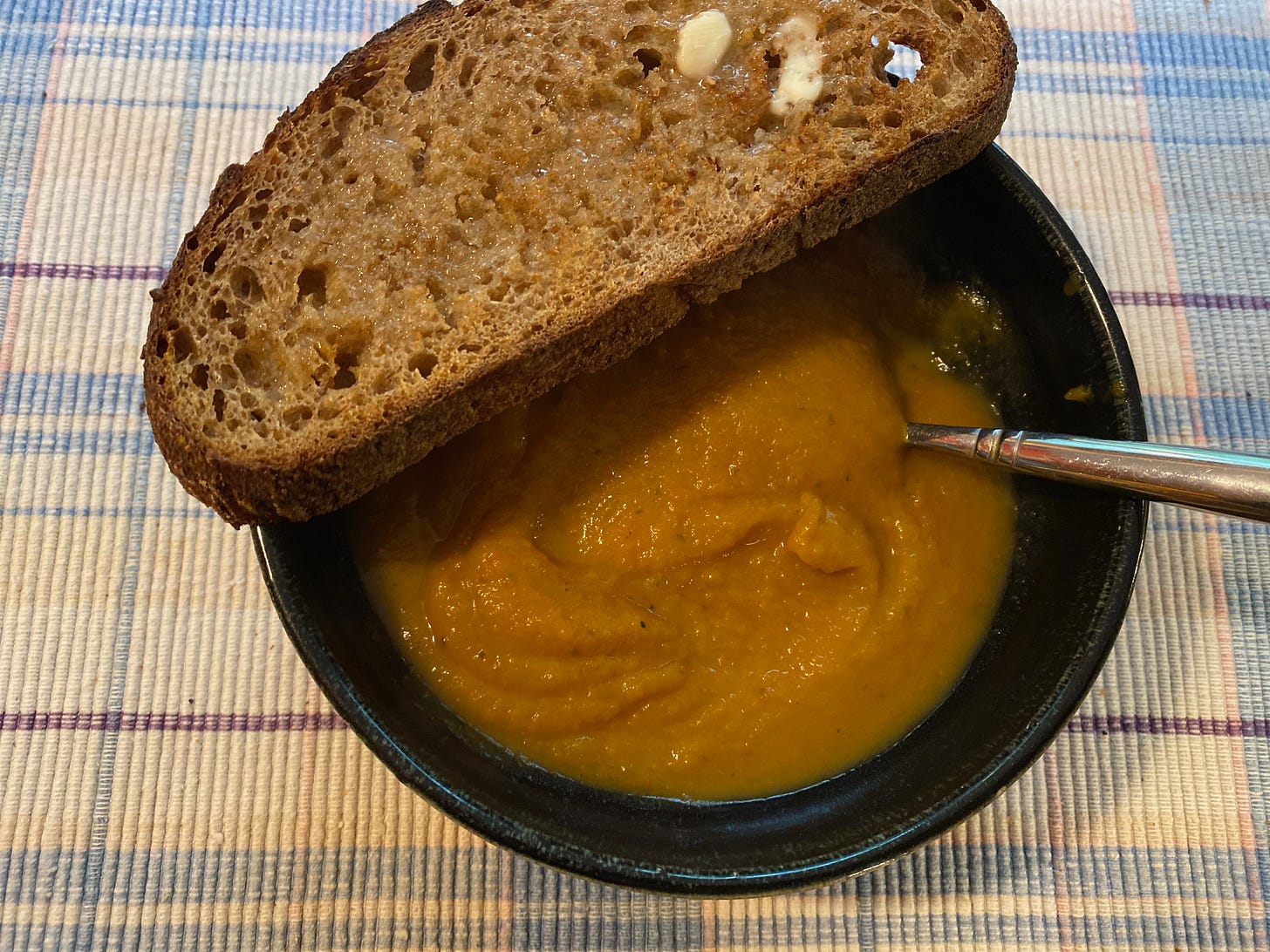 A ceramic bowl or sweet potato soup sits on a blue and white placemat. A slice of buttered toast sits on the edge of the bowl.