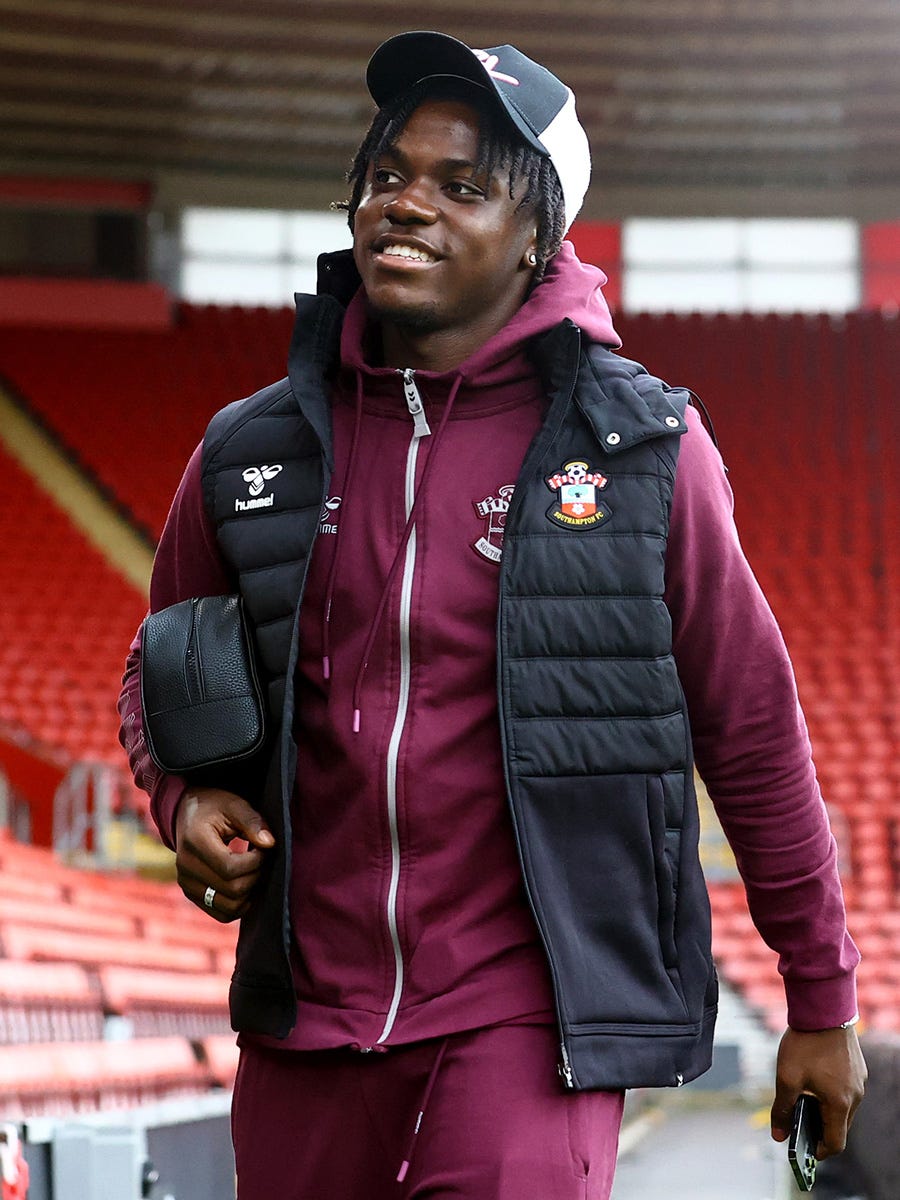 Romeo Lavia smiles and looks to his right as he walks through St Mary's on his way to the tunnel. The midfielder is wearing a purple Saints-branded tracksuit and black gilet, with a black-and-white baseball cap, while carrying his phone in his left hand and washbag under his right arm.
