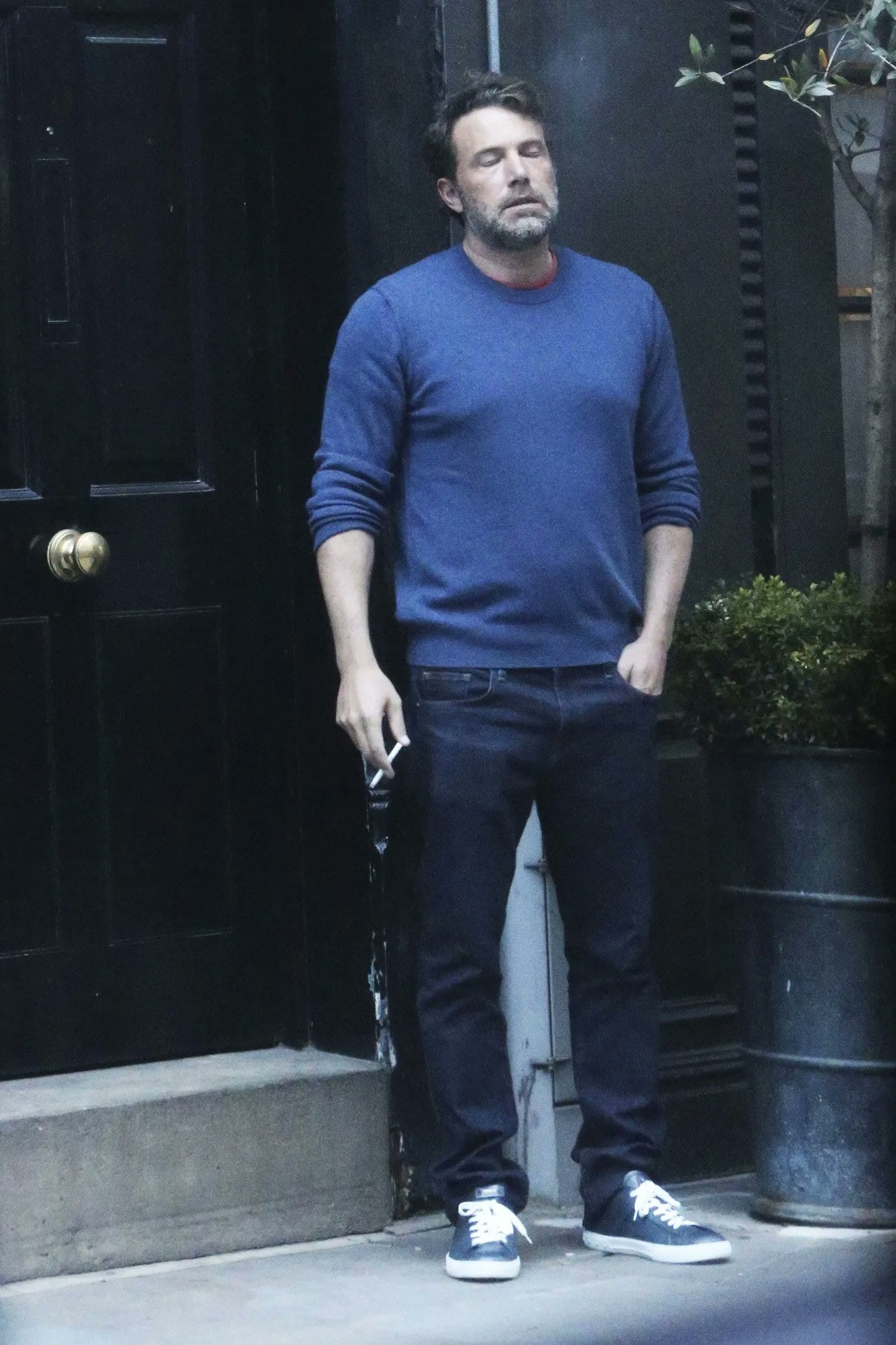 A picture of actor Ben Affleck wearing jeans and a blue sweater while standing outside a building smoking a cigarette and looking like he is done with life.