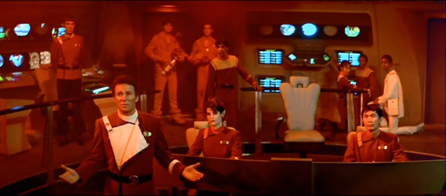 Frame from Star Trek movie showing the bridge with red alert lights on and crew standing as Kirk speaks with Khan telling him the bridge is smashed and computers inoperable.