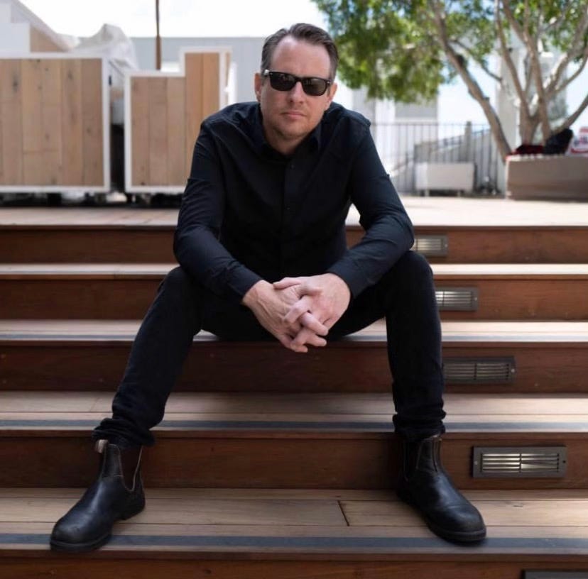A man dressed in all black (it's John Sparrow of the Violent Femmes! That's who this article is about!) and wearing sunglasses sits on some kind of outdoor wooden stairs. It's sunny and there's a tree in the background. I love it.