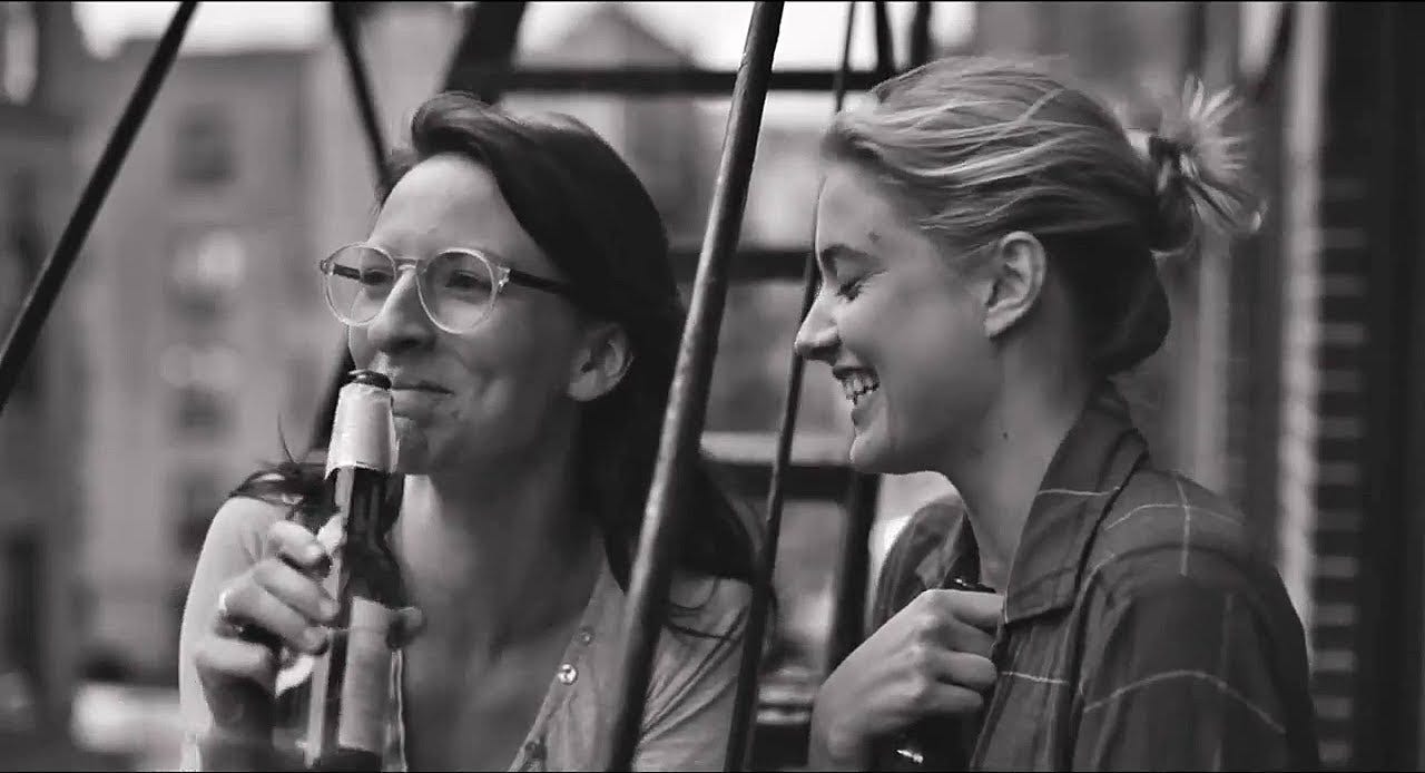 Descrição: imagem do filme Frances Ha em que duas personagens mulheres riem enquanto bebem cerveja em uma escada ao ar livre.