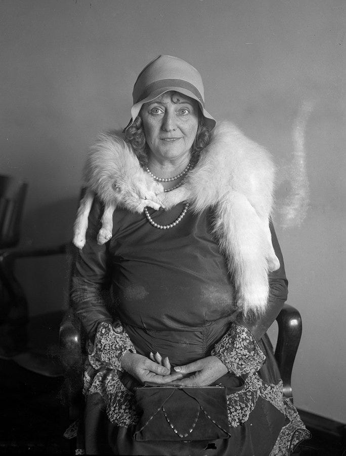 A black and white photograph of Dolly around the time of the trial. She is middle aged, sat on a chair, and looks every bit your 1930s glamour puss. She is wearing a fox fur around her neck, a long dress with laced sleeve details, and a bucket hat. Her hair is curled & she has her handbag on her knees. This is not someone you'd think would have a gimp stashed in the attic. 