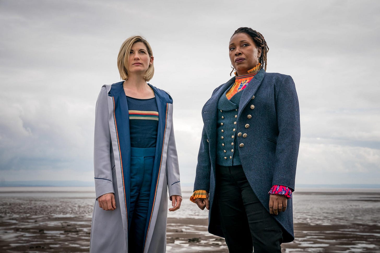 This image shows the Thirteenth Doctor (played by Jodie Whittaker) on the left and Ruth Clayton--aka the Fugitive Doctor (played by Jo Martin)--on the right. They stand on a beach, with a sliver of sea and large clouds visible in the background.