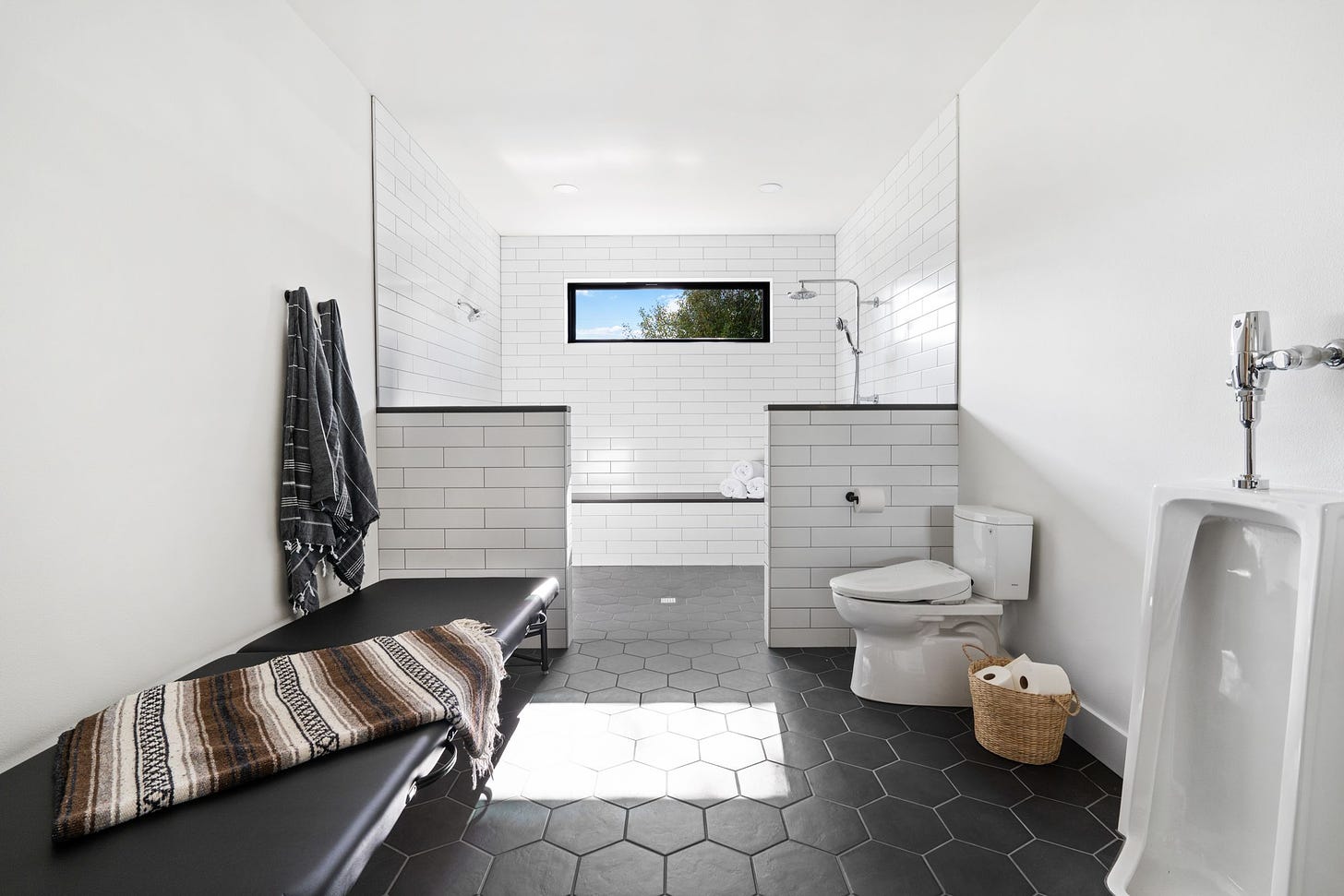 White subway tiles panel a roll-in shower. A bench holds rolled white towels inn the background while a window shows a pop of blue sky. In the foreground, a black mat bench topped with a brown southwest style blanket. On the right, a toilet and a urinal with plenty of open space. 