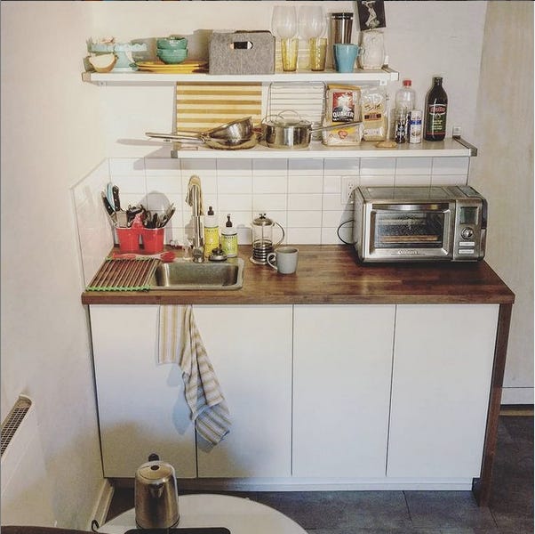 A counter top the width of two cabinets with two shelves above. There's a tiny bar sink, a toaster oven, a kettle, and a hot plate. There are two shelves above holding dishes, pans, and food. The cabinets hold a mini fridge and the water heater.