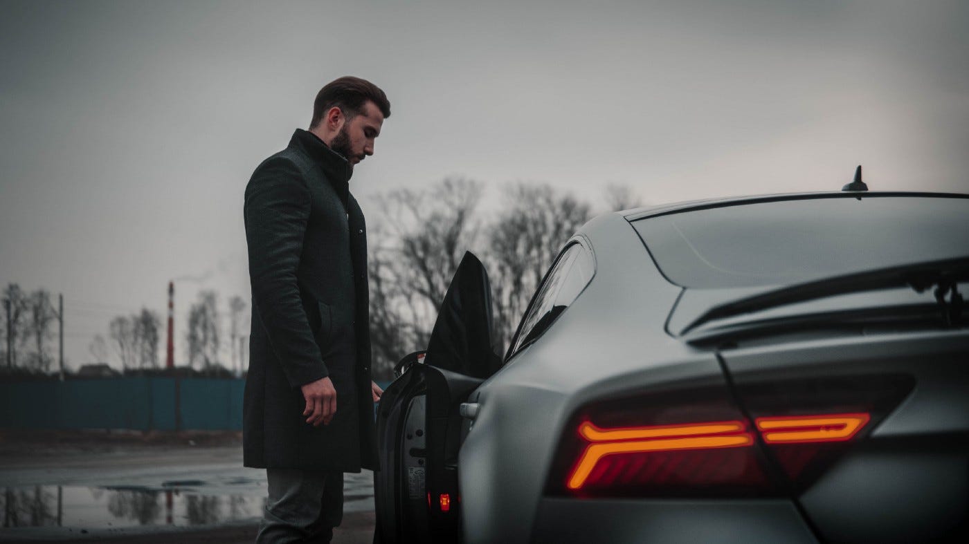 Man in an overcoat standing next to a supercar