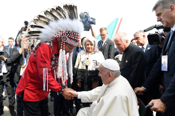 An elderly white man dressed in a white coat and wearing a white cap sits on a wheelchair and shakes the hand of another elderly man who is standing and wearing a traditional headdress and a red traditional shirt decorated with beading and tassels. Behind them stand many onlookers and media filming and taking pictures.