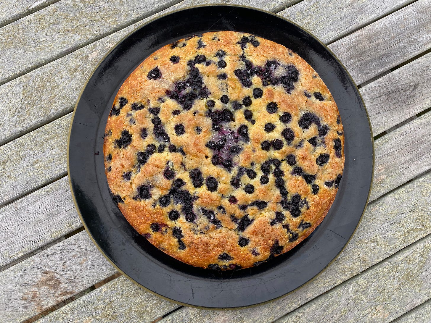 A round golden brown cake full of blueberries on a black platter.