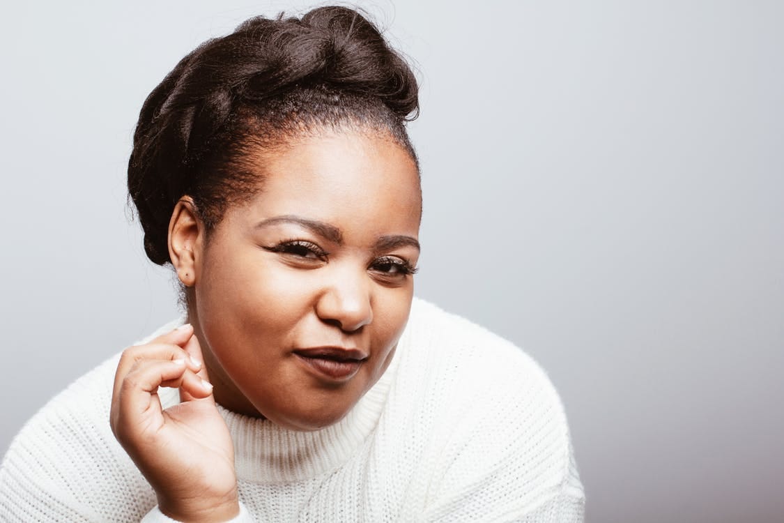 Positive plus size African American female wearing cozy white knitted sweater standing on gray background in studio and looking at camera with half closed eyes