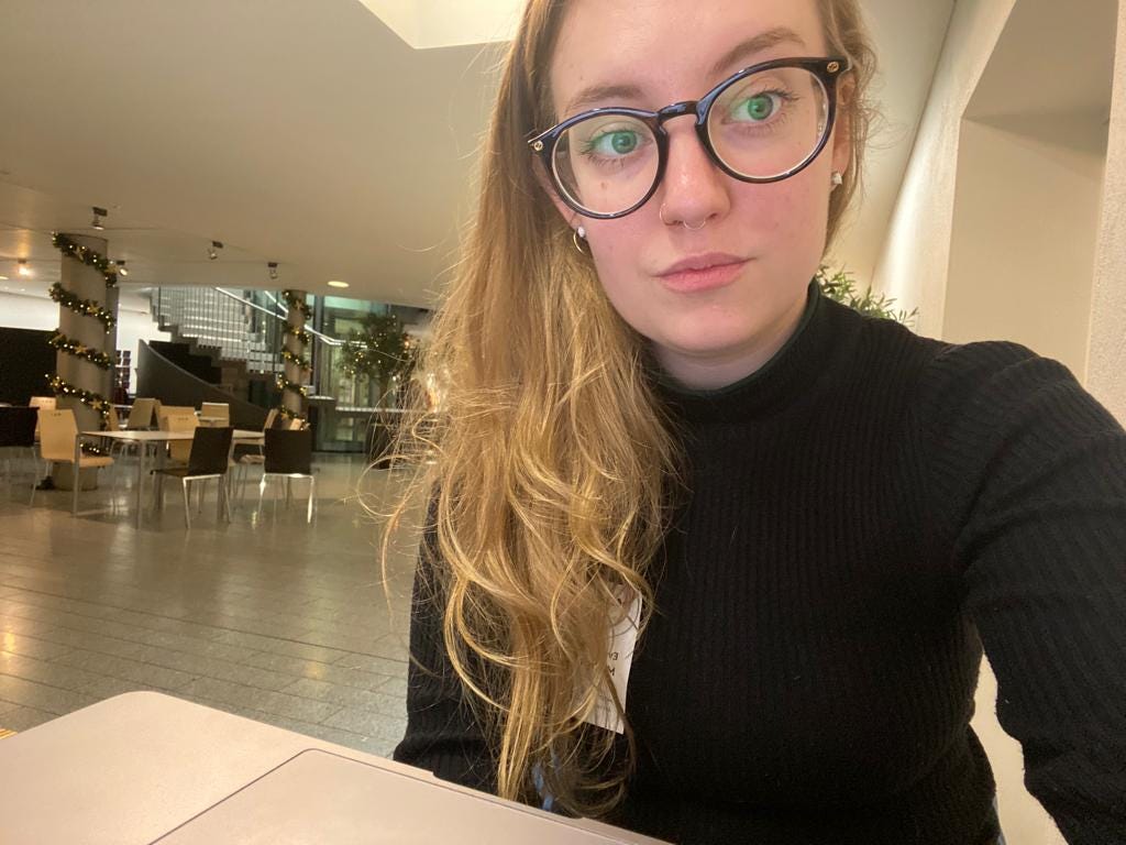a female-bodied person with long, wavy hair, wearing a black jumper, bold framed glasses and a small ring in her nostril and septum. she sits in Café in the Crypt in Trafalgar Square.