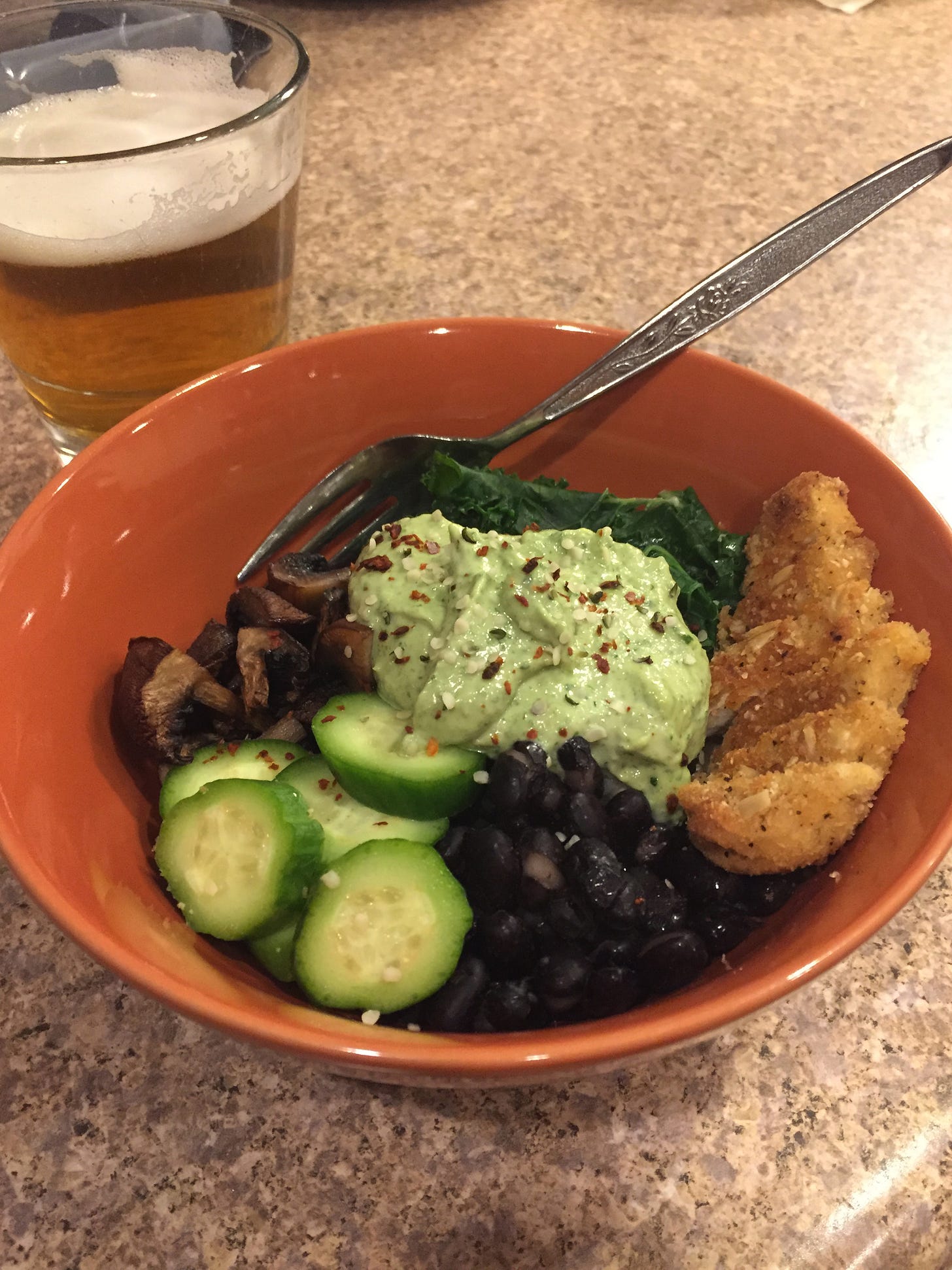 In an orange bowl, small piles of black beans, cucumber, roast mushrooms, kale, and a chopped chick'n tender sit on top of of brown rice. A blob of green goddess dressing is in the centre, with hemp seeds and chili flakes flecked on top.