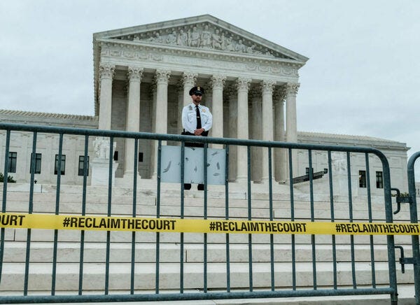 A guard stands atop the steps leading to the Supreme Court, which looms up behind him. At the foot of the stairs, in the foreground, we see a barrier, lined by yellow tape upon which is printed the legend "#RECLAIMTHECOURT"